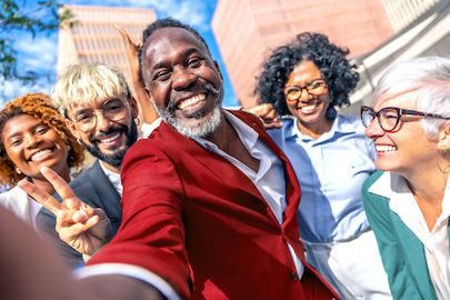 Oameni de diferite vârste, rase și sexe zâmbind larg în fața unei clădiri